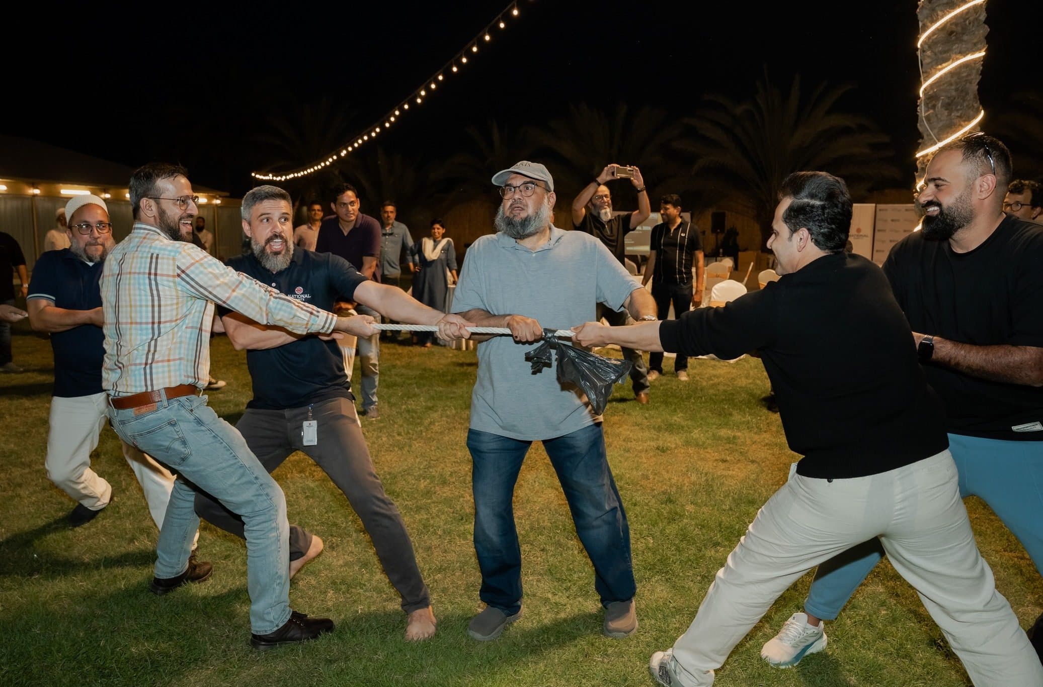 Build&Bond - BMG Social 2026: Men playing tug of war on grass at night under string lights