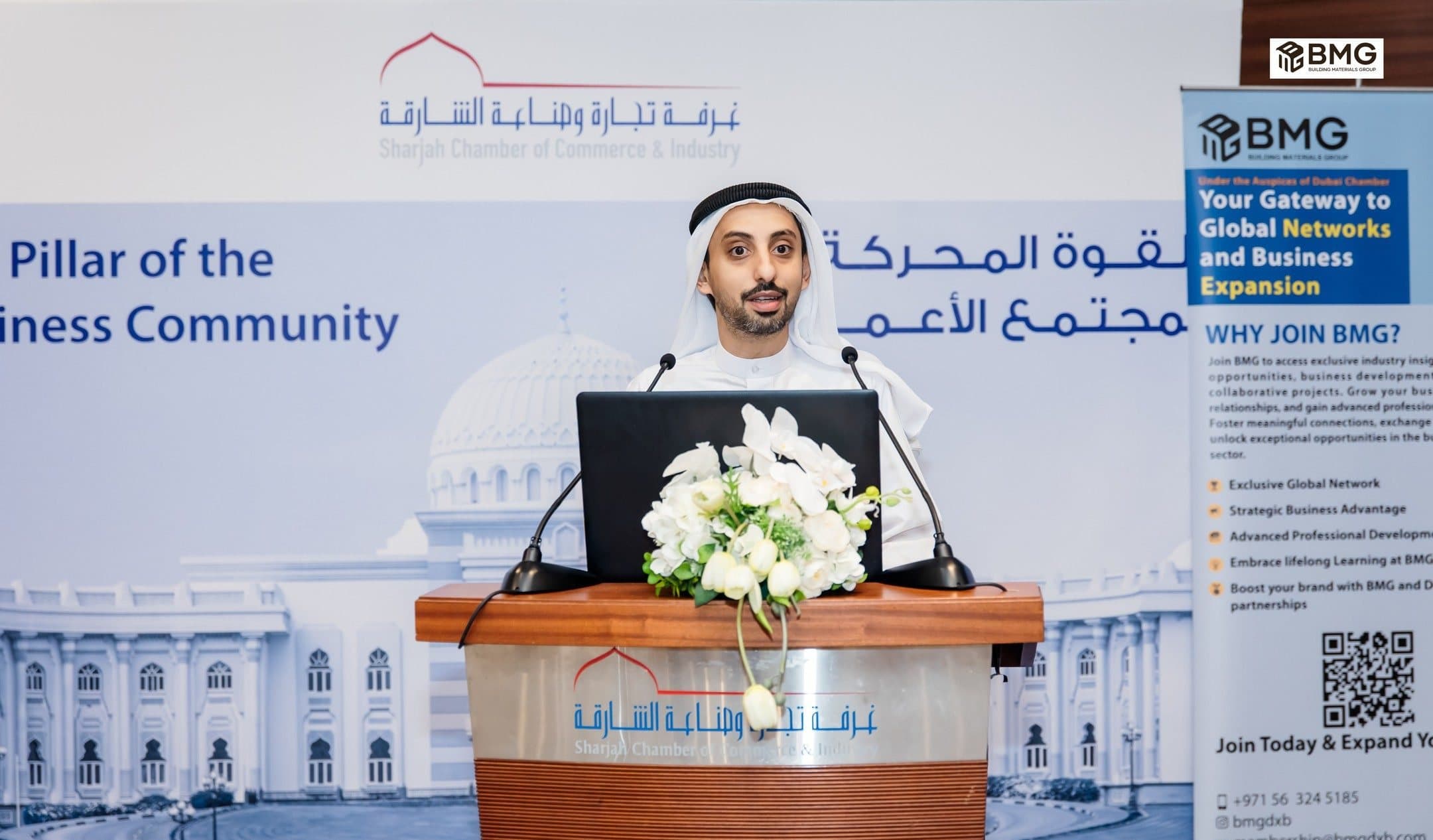 Building Materials Forum: Emirati speaker at a Sharjah Chamber podium with flowers, BMG roll-up banner on the right