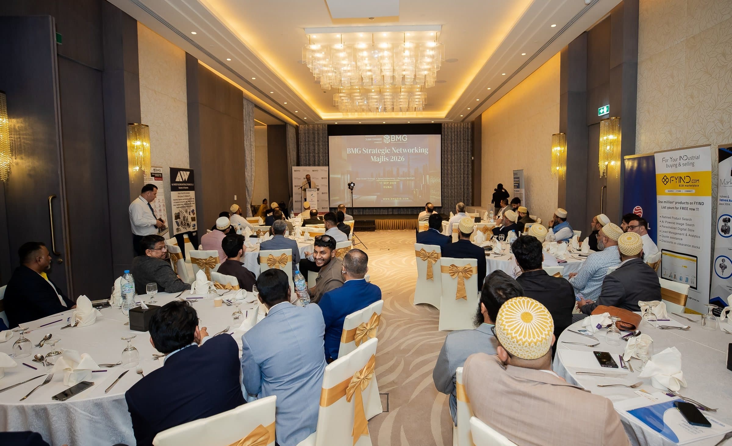 Wide ballroom view with guests at round tables facing a screen, chandeliers above
