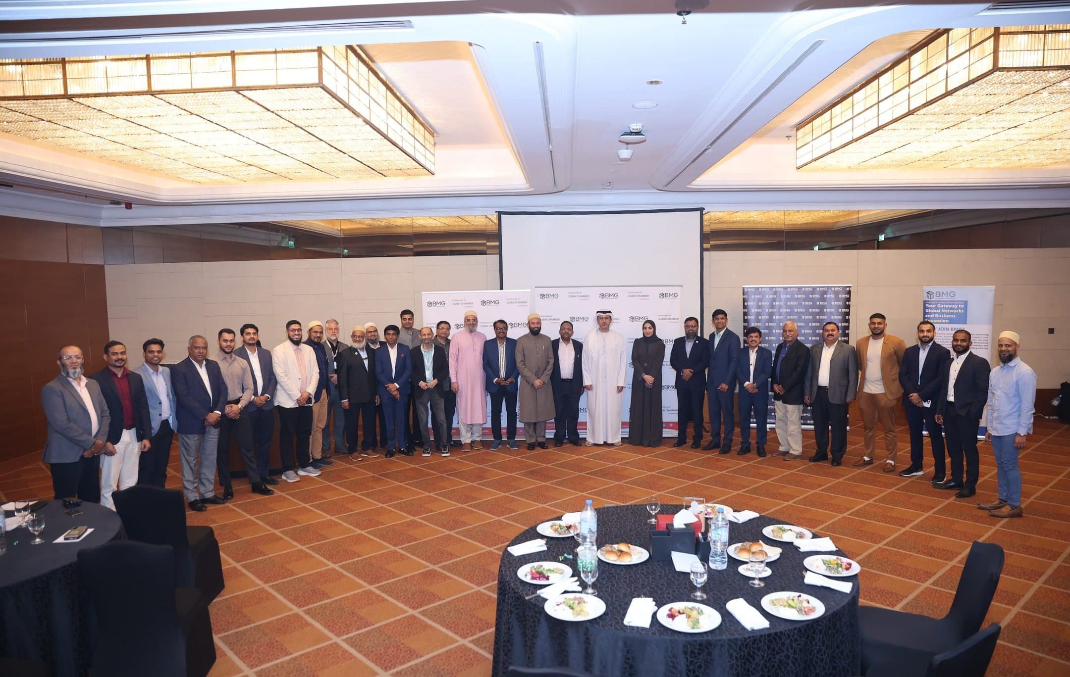 Guests standing in a wide semicircle in a hotel hall around a set dinner table