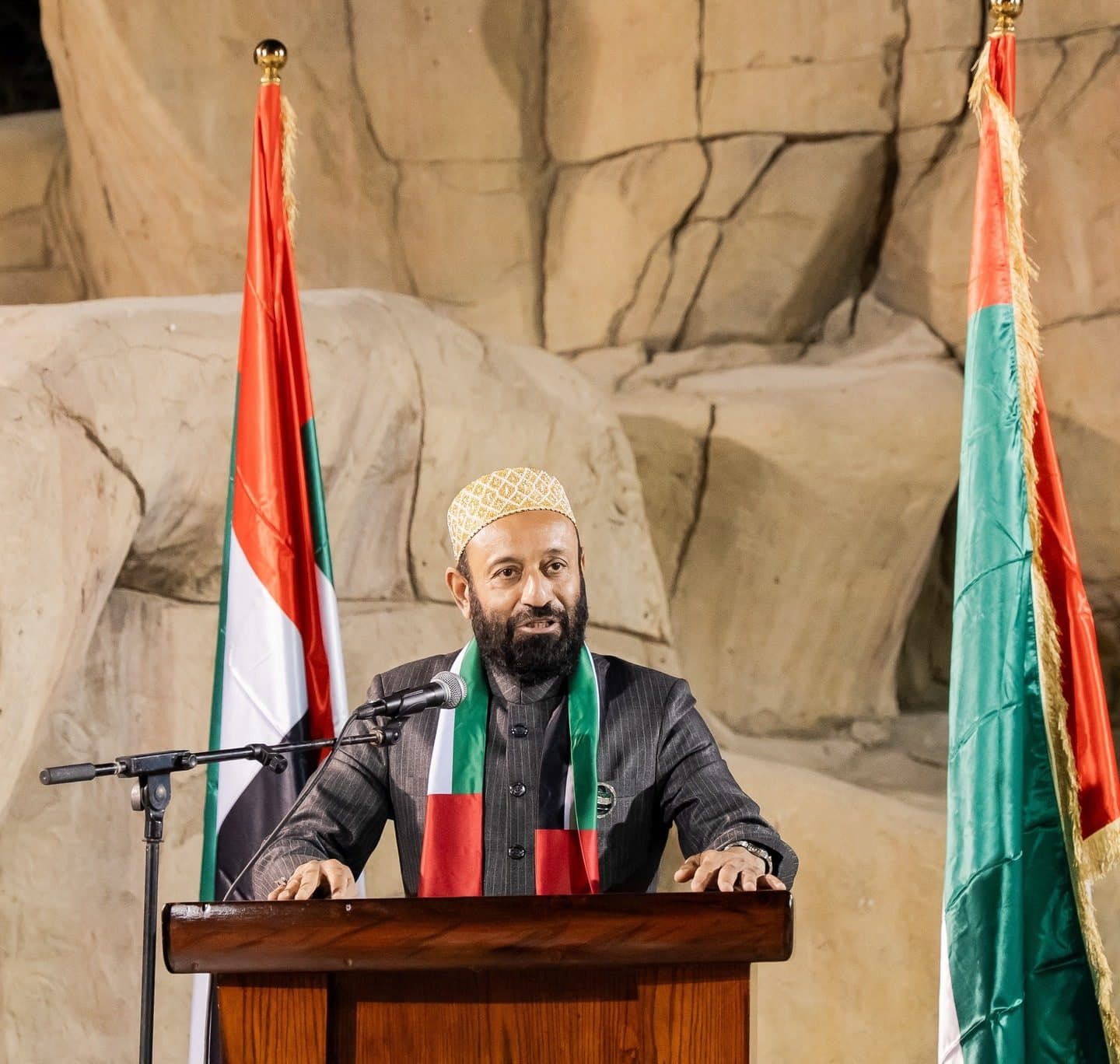 54th UAE National Day Celebration: Speaker at a wooden podium between two UAE flags, rock wall backdrop
