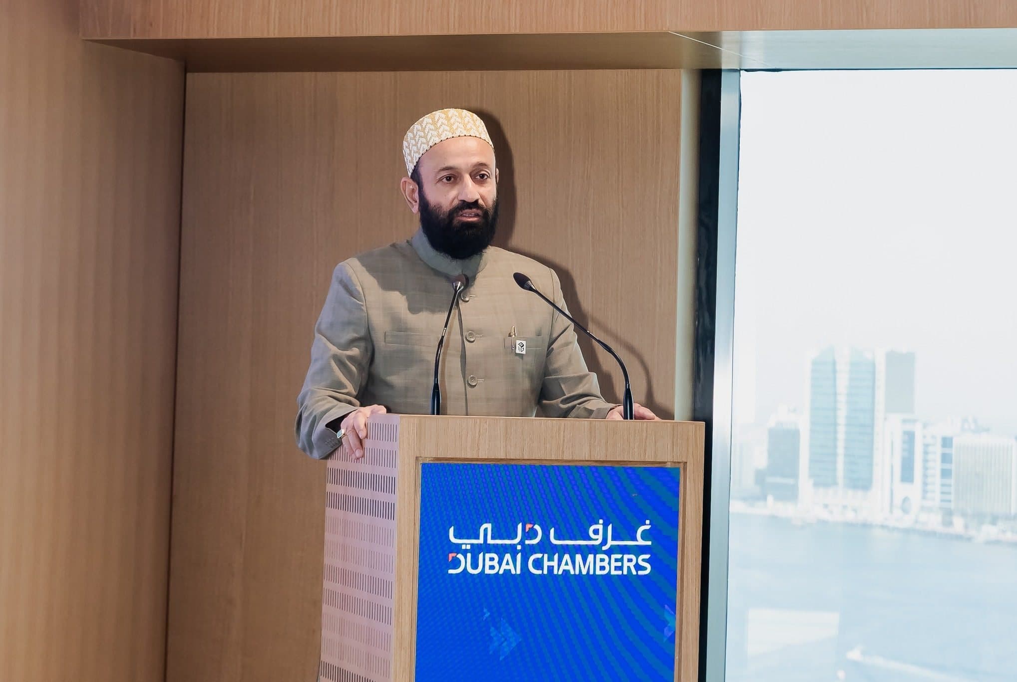 Annual General Meeting 2025: Man in a cap speaking at a Dubai Chambers podium beside a window