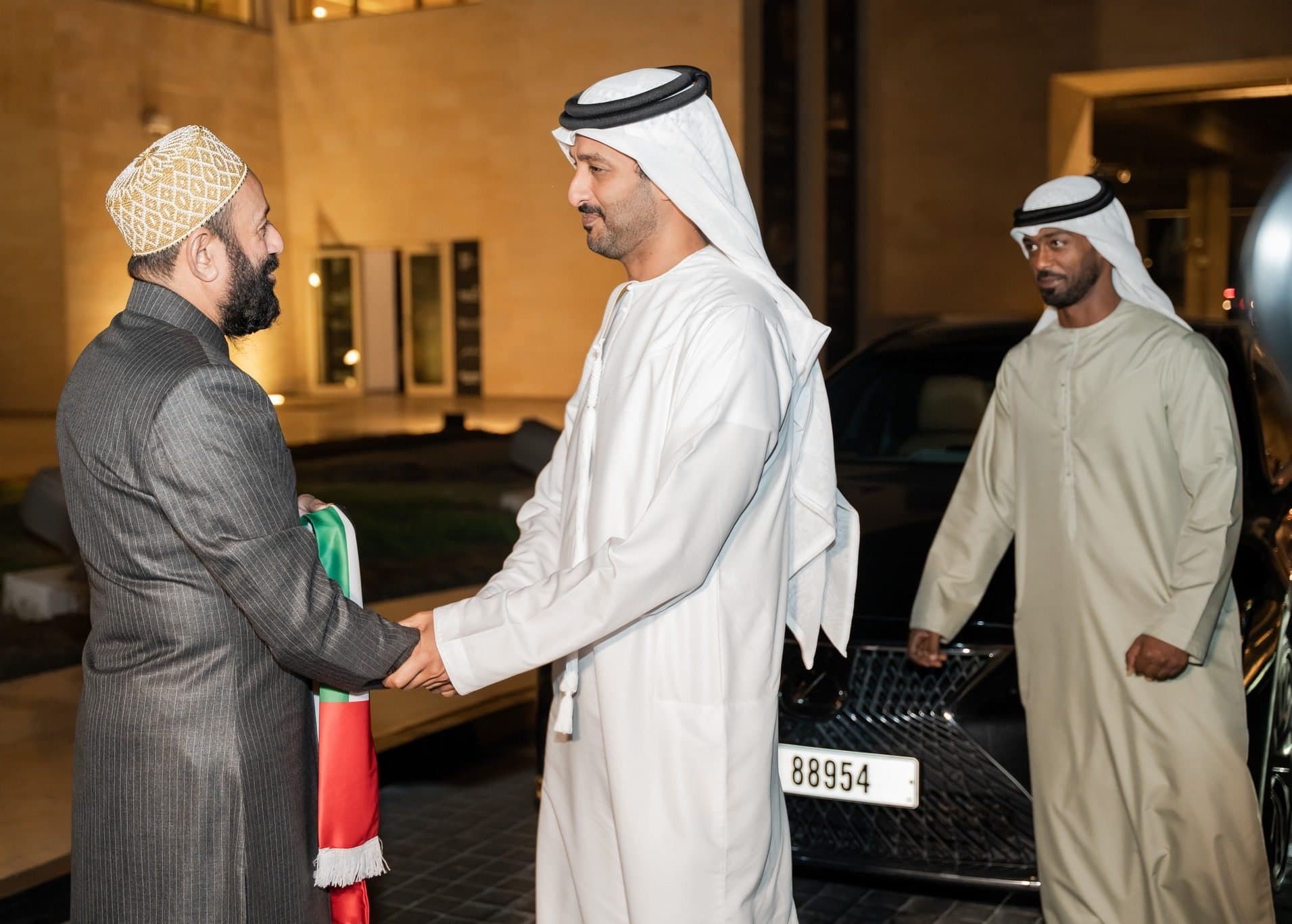 Man in a cap greeting an Emirati guest wearing a UAE flag scarf at an evening arrival