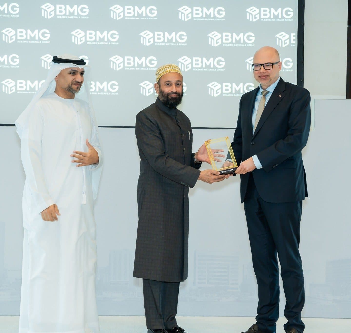 Three men presenting a trophy in front of a BMG logo wall, one Emirati in kandura