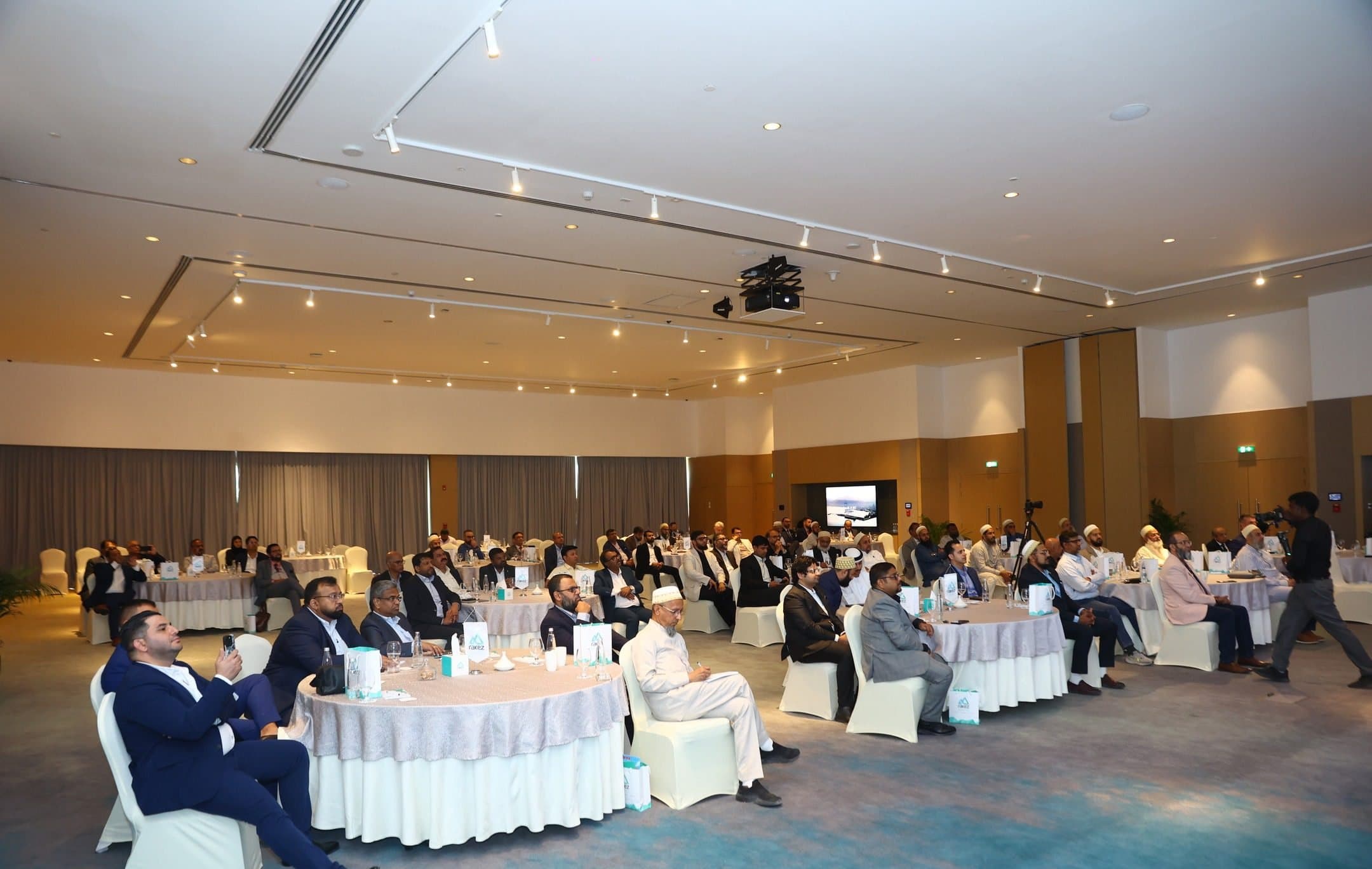 Wide view of a hotel ballroom with guests seated at round tables facing a screen