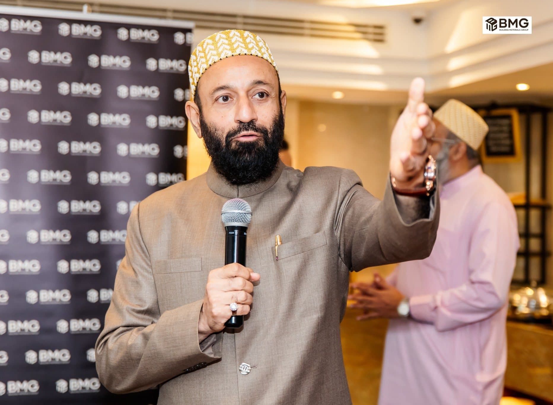 Man in a cap speaking into a microphone and pointing, BMG logo wall behind