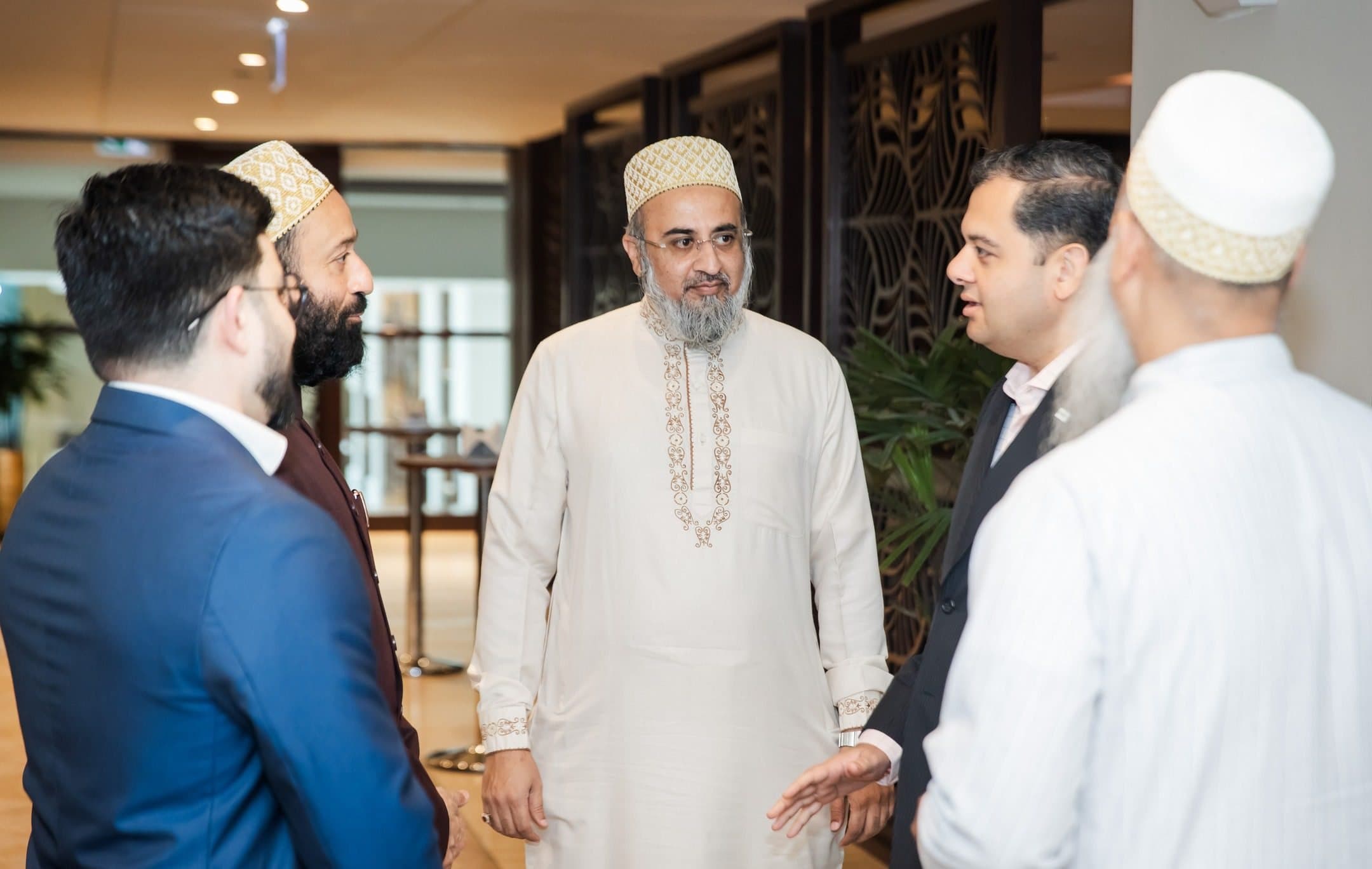 Four men in conversation, one in a white embroidered kurta and cap