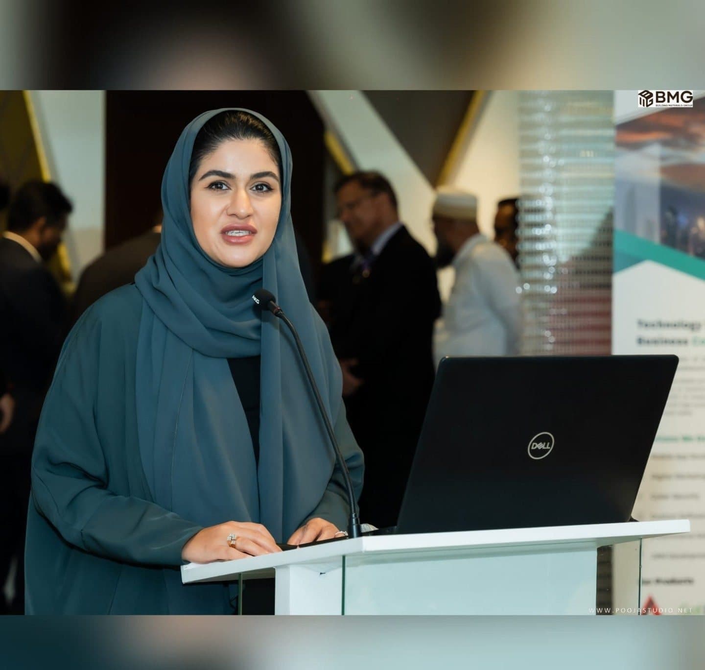 Woman in a green abaya and hijab speaking at a white podium with a laptop