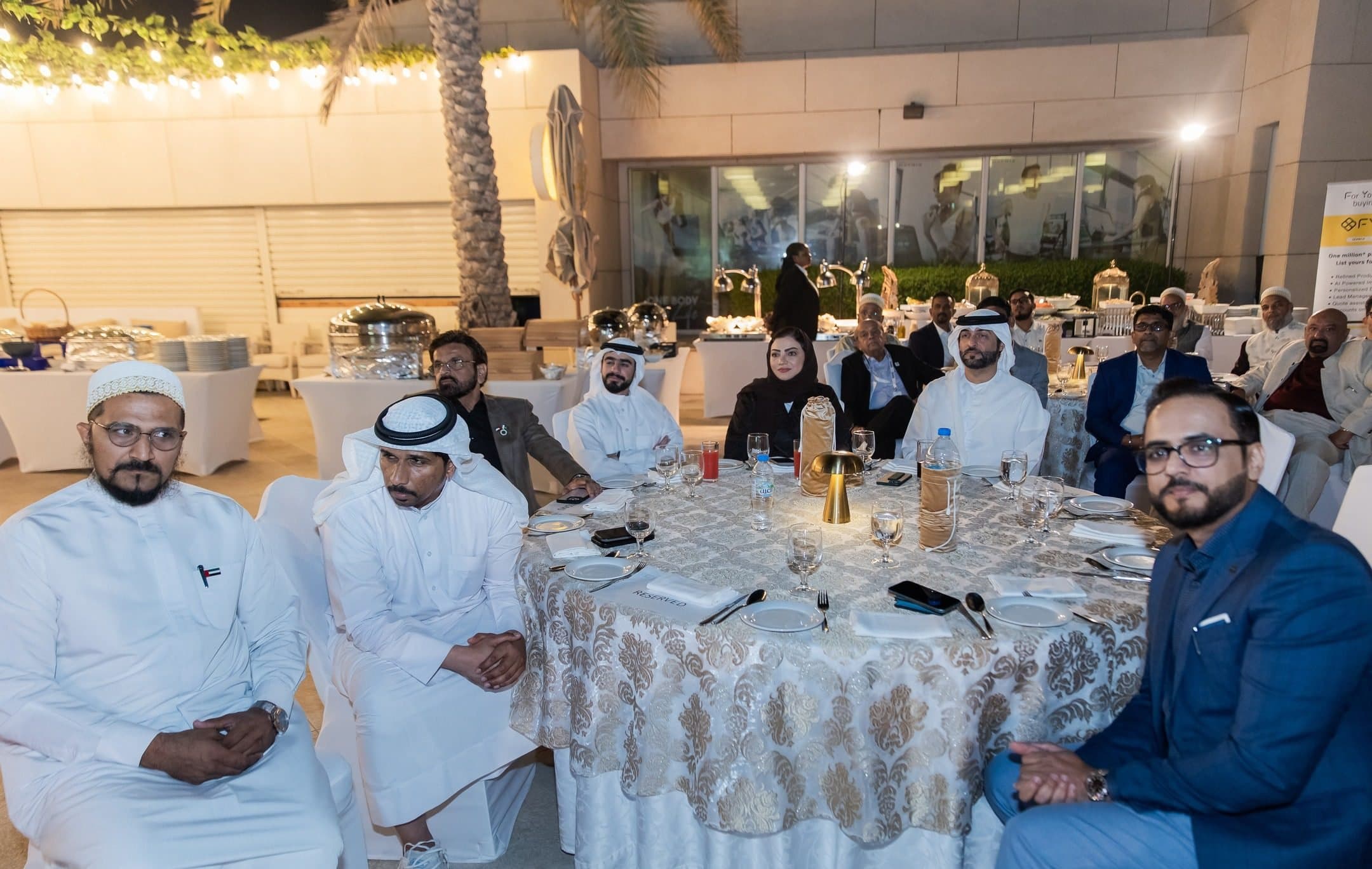 Guests seated around a round dinner table outdoors at night, some in kandura