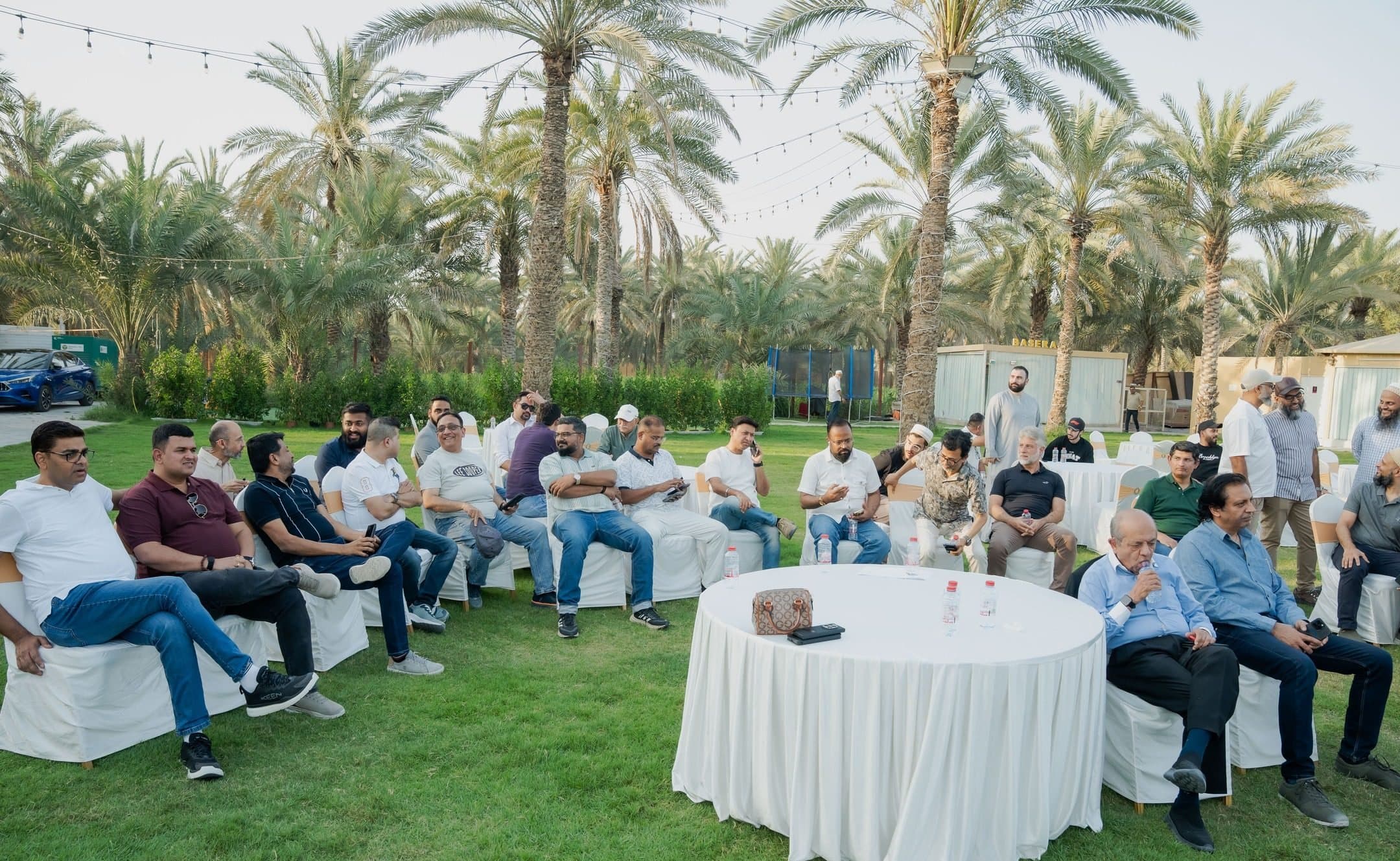 Members sitting in a circle of chairs on a lawn under palm trees