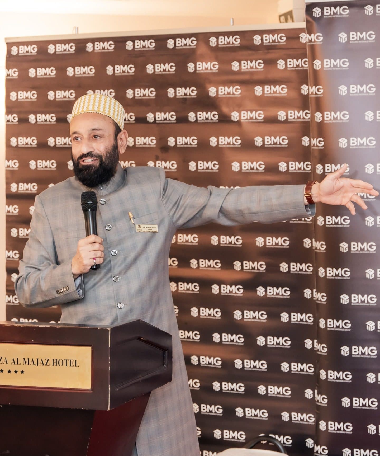 Speaker in a cap gesturing at a hotel podium in front of a BMG logo wall