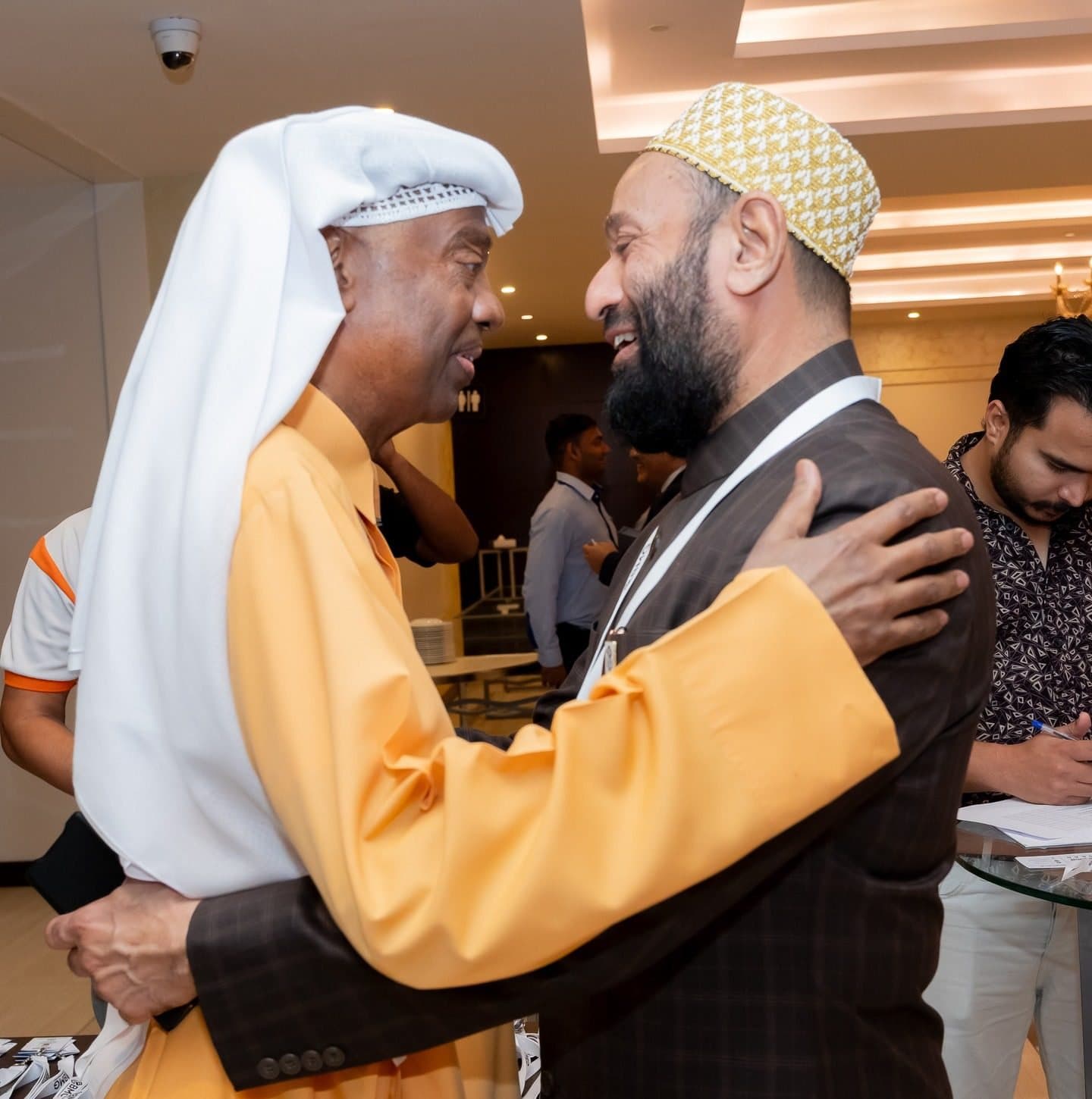 Man in a cap embracing a guest in a yellow robe and white headdress