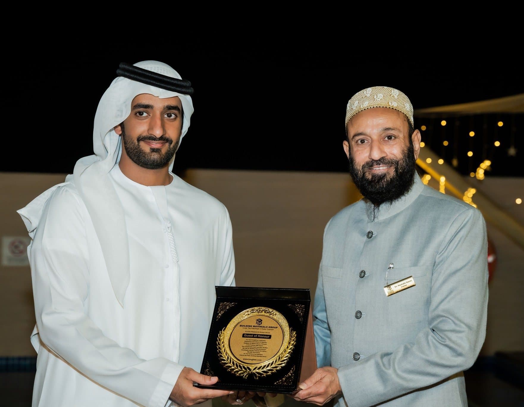 Two men holding a gold plaque at a night rooftop event with string lights